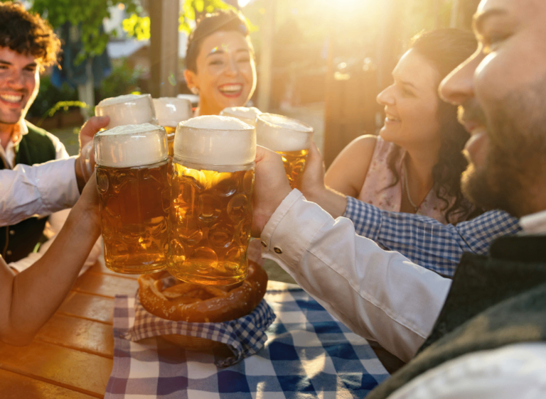 Eine Gruppe von Menschen im Freien an einem Tisch, die große Gläser Bier halten und lächeln. Ein Brezel liegt auf einem blau-weiß karierten Tuch.