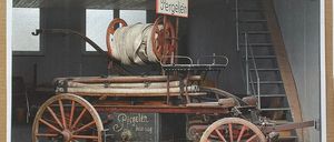 Ein altes Feuerwehrauto steht drinnen mit einem Schild, auf dem 'Löschzug der Feuerwehr Pergelen' steht. Das Auto ist rostig und hat ein Rad auf jeder Seite. Es hat eine Leiter auf der rechten Seite. Das Gebäude hat Fenster und eine Treppe.