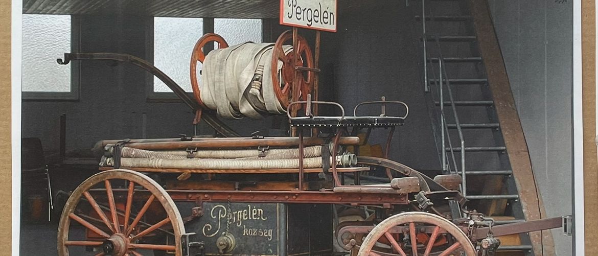 Ein altes Feuerwehrauto steht drinnen mit einem Schild, auf dem 'Löschzug der Feuerwehr Pergelen' steht. Das Auto ist rostig und hat ein Rad auf jeder Seite. Es hat eine Leiter auf der rechten Seite. Das Gebäude hat Fenster und eine Treppe.