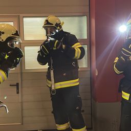 Drei Feuerwehrleute in voller Ausrüstung, einschließlich Helme und Taschenlampen, stehen in der Nähe einer Tür mit Glasfenstern und einem Griff. Sie bereiten sich wahrscheinlich auf eine Übung vor.