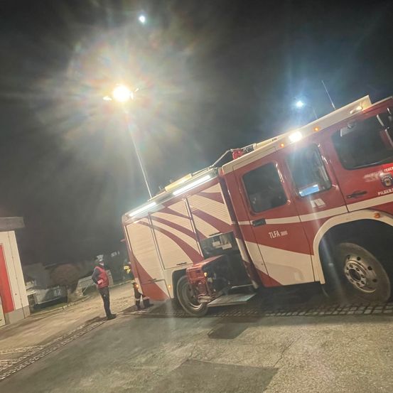Ein Feuerwehrfahrzeug ist nachts in der Nähe eines Gebäudes geparkt, wobei eine Person in der Nähe steht. Das Fahrzeug hat seine Lichter eingeschaltet und wird von einer Straßenlaterne beleuchtet.