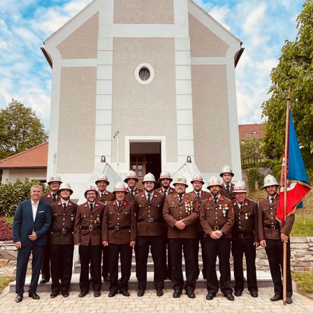 Eine Gruppe Feuerwehrleute in Uniform steht vor einer Kirche mit einer Flagge an einem sonnigen Tag.