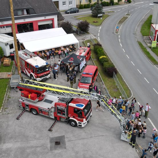 Eine Vogelperspektive einer Feuerwache mit mehreren geparkten Feuerwehrautos, einer Gruppe von Menschen, die herumstehen, und einer Leiter, die von einem der Trucks ausgestreckt ist.