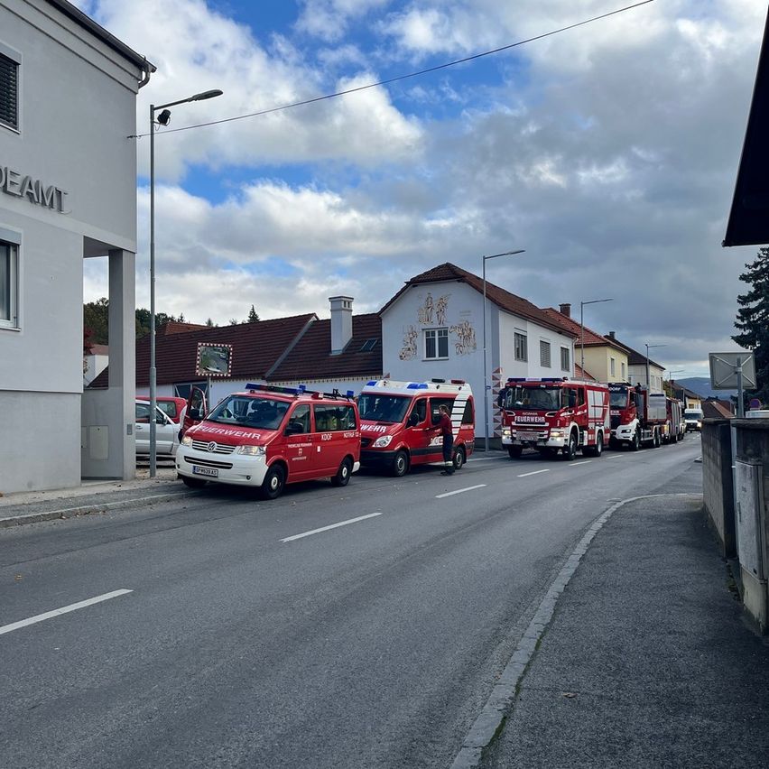Mehrere rote Feuerwehrfahrzeuge sind am Straßenrand geparkt, in der Nähe von Gebäuden und Häusern. Der Himmel ist blau mit Wolken.