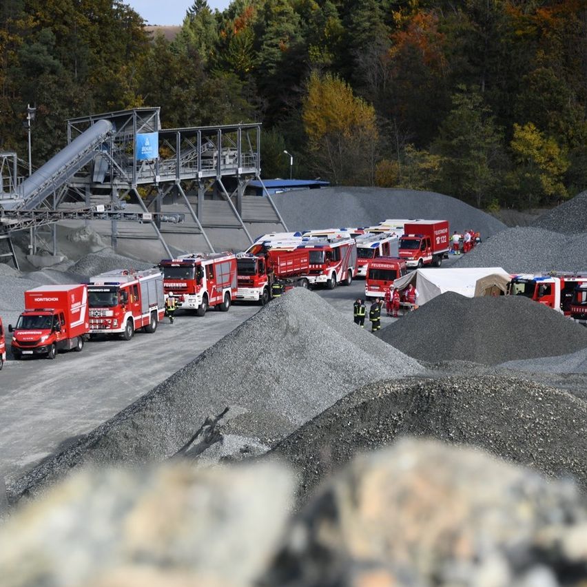 Mehrere rote Feuerwehrfahrzeuge sind auf einer Kiesoberfläche geparkt, mit mehreren Haufen Kies in der Umgebung. Eine Gruppe von Feuerwehrleuten ist in der Nähe der Fahrzeuge versammelt. Im Hintergrund befindet sich ein Zelt.