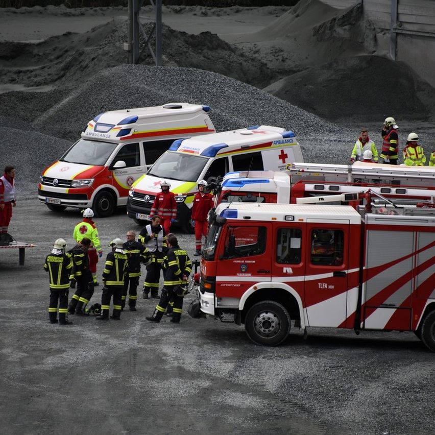 Feuerwehrleute und Sanitäter stehen um ein Notfallfahrzeug mit Leiter. Zwei Rettungswagen sind im Hintergrund in der Nähe eines Schotterhaufens geparkt.