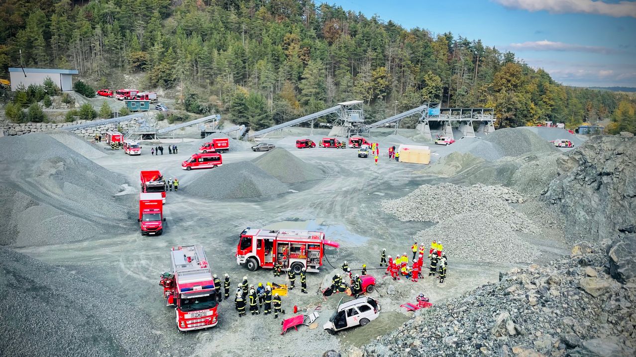 Eine Draufsicht auf eine Mine zeigt mehrere rote Feuerwehrwagen und Rettungsfahrzeuge. Viele Menschen, die reflektierende Westen tragen, stehen um einen beschädigten weißen SUV herum. Die Szenerie ist vor dem Hintergrund von Kies und Bergbaugeräten gelegen.