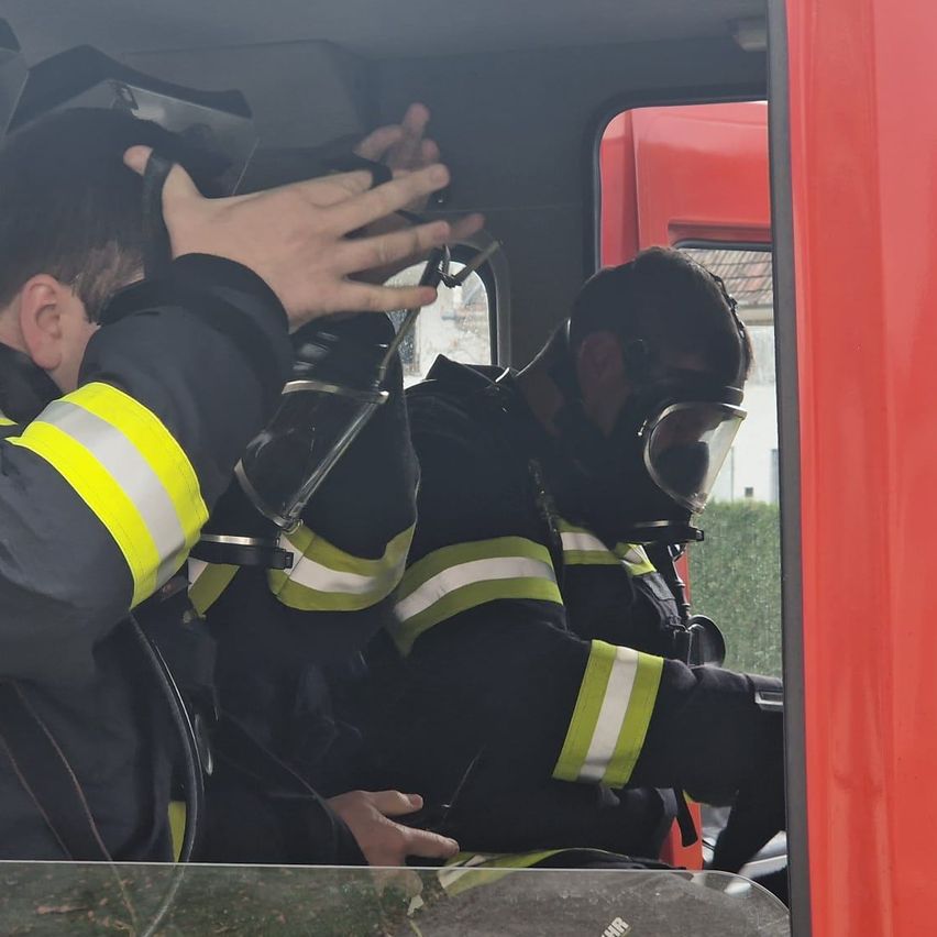 Two firefighters in full gear are inside a fire truck, one with a gas mask and the other adjusting his helmet. The truck's door is open.