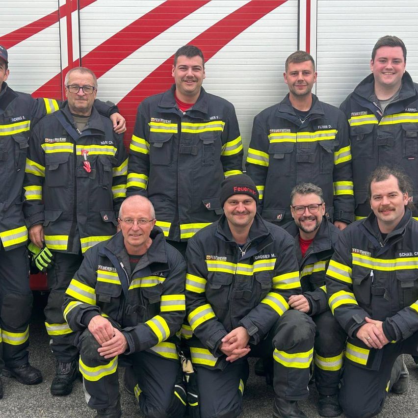 A group of firefighters in uniform stand and crouch in front of a red and white fire truck. They are all smiling.