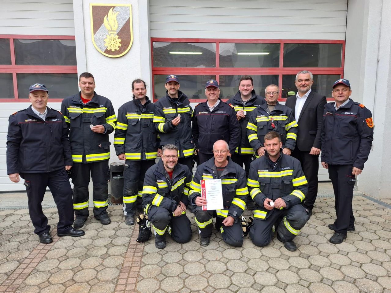 A group of firefighters pose outside a fire station with one holding a certificate.