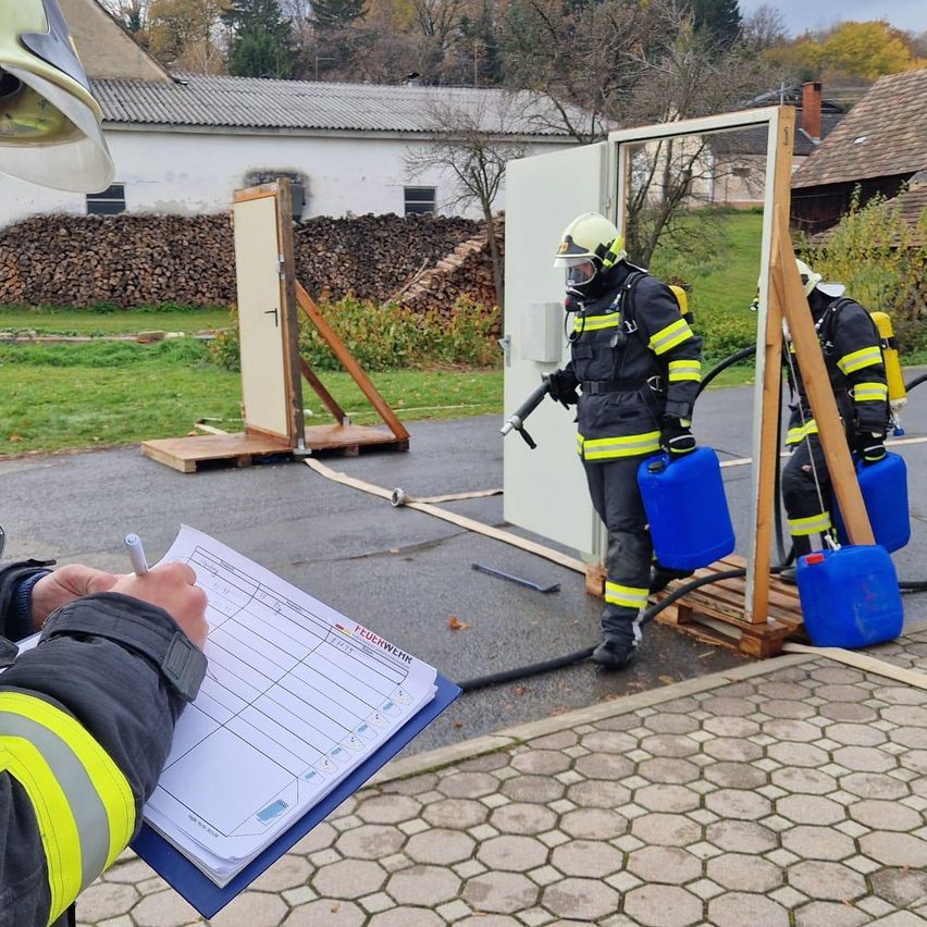 Two firefighters are practicing opening a door and spraying water, while another is holding a hose and a container. A person in a helmet is observing from the side.