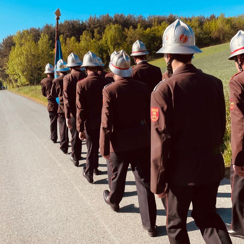 Eine Gruppe von Feuerwehrleuten in Uniform marschiert auf einer Straße, mit Bäumen und einer Flagge im Hintergrund.