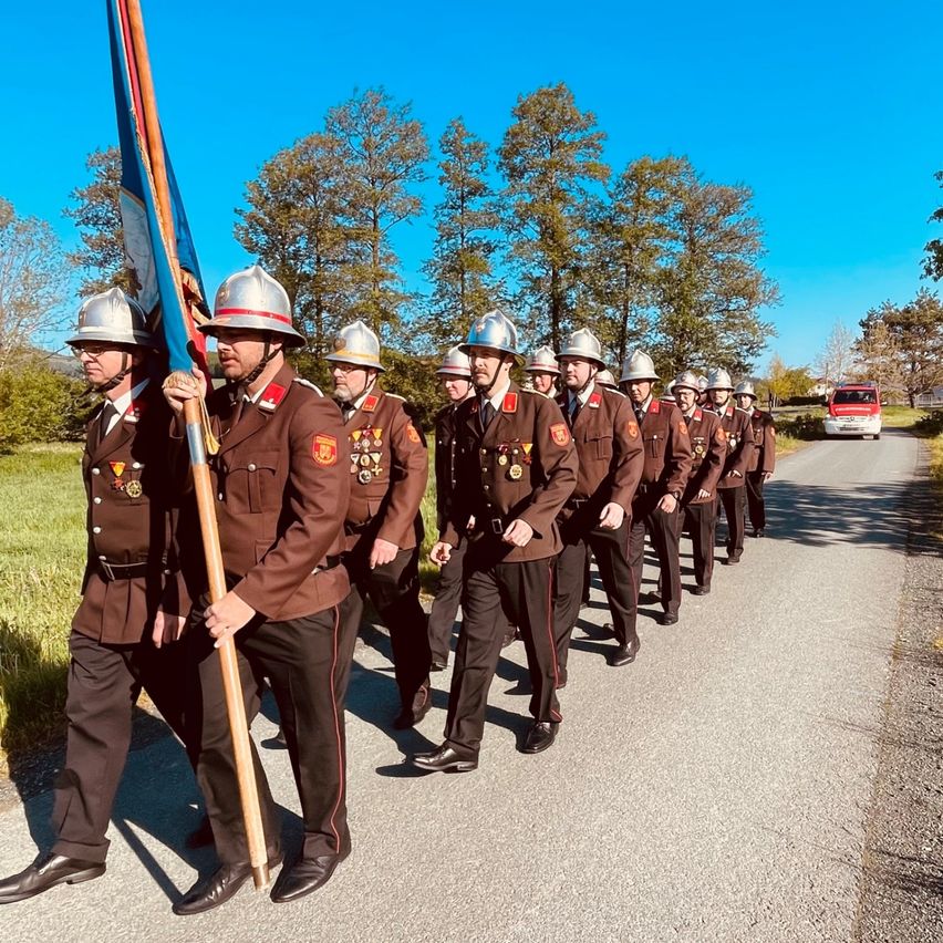 Eine Gruppe von Feuerwehrleuten marschiert auf einer Straße, trägt Uniformen und trägt eine Fahne. Die Straße ist von Gras und Bäumen umgeben, und ein rotes Fahrzeug fährt vorbei.
