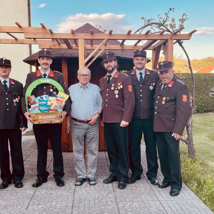 Fünf Männer in Uniformen posieren für ein Foto, einer hält einen Korb mit Snacks. Sie stehen auf einem Ziegelweg, mit einem hölzernen Pavillon im Hintergrund.