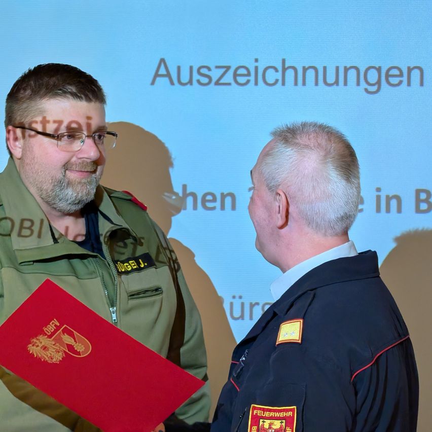 A man in a military uniform is presenting a red folder to another man in a blue uniform. Behind them is a blue screen with white text.