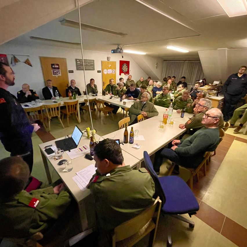 A group of individuals in military uniforms are seated around tables in a room. One person stands at the front, possibly giving a presentation. Bottles and cups are on the tables, and a projector is mounted on the ceiling.