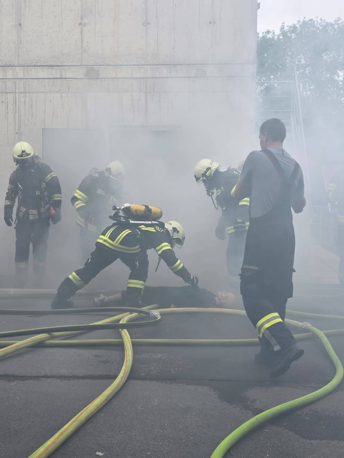 Feuerwehrleute in voller Ausrüstung führen eine Rettung in einer rauchigen Umgebung durch. Ein Feuerwehrmann liegt am Boden, während andere beobachten.