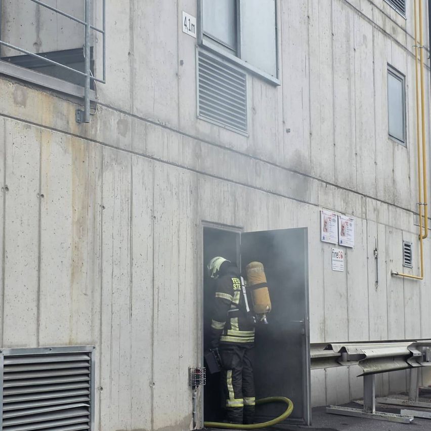 Ein Feuerwehrmann verlässt ein Gebäude mit einem Sauerstofftank, Rauch erfüllt die Luft. Das Gebäude hat Betonwände und Metallgeländer.