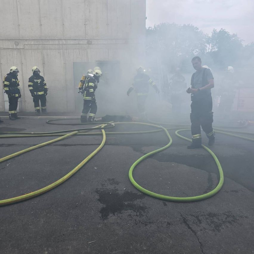 Feuerwehrleute stehen um eine offene Gebäudetür herum. Ihnen kommt Rauch aus dem Gebäude. Ein Mann steht vor ihnen.