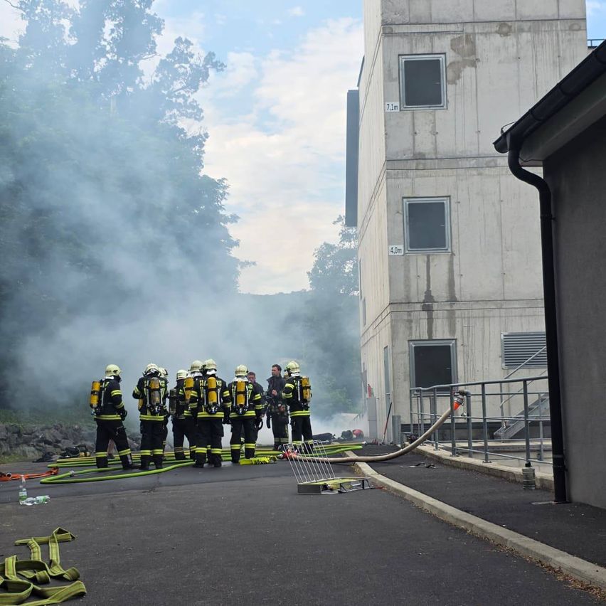 Eine Gruppe von Feuerwehrleuten steht vor einem Gebäude, aus dem Rauch kommt. Sie tragen alle Helme und Rucksäcke.