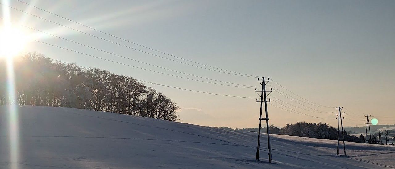 Ein Strommast steht allein auf einem verschneiten Hügel mit laublosen Bäumen im Hintergrund.