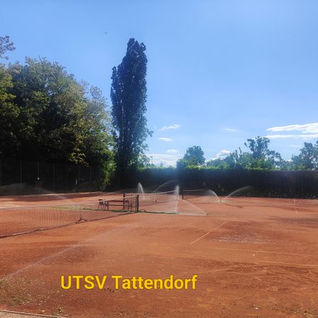 Ein Tennisplatz mit einer Bank und einem Netz. Der Boden ist mit Erde bedeckt. Bäume stehen hinter dem Zaun. Wassersprüher sind auf dem Boden. Der Text UTSV Tattendorf befindet sich unten rechts.