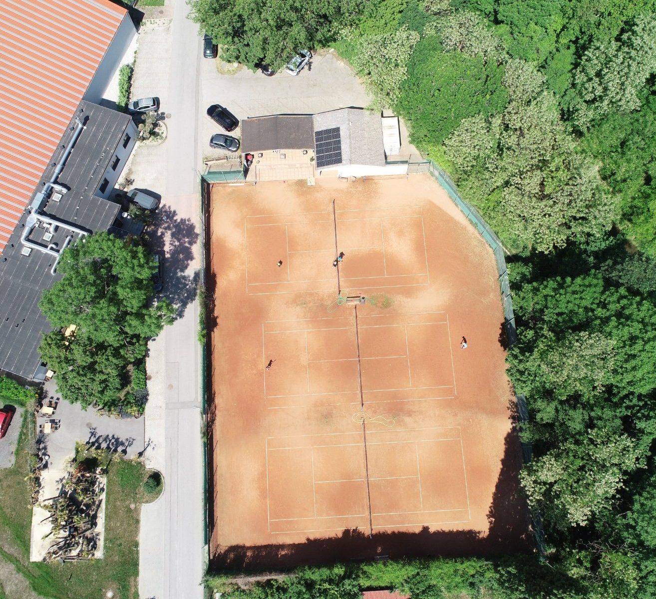 Luftaufnahme eines Tennisplatzes umgeben von Bäumen mit einem Gebäude und geparkten Autos.
