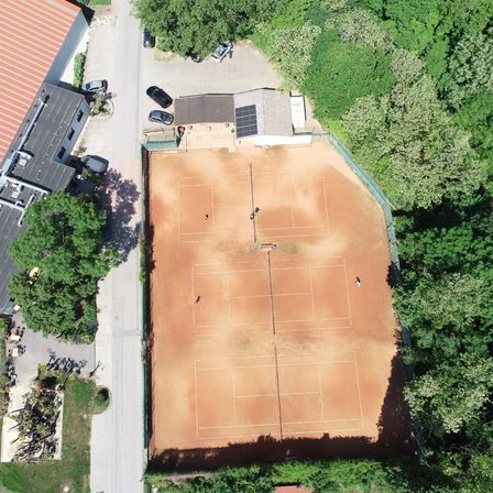 Luftaufnahme eines Tennisplatzes umgeben von Bäumen mit einem Gebäude und geparkten Autos.
