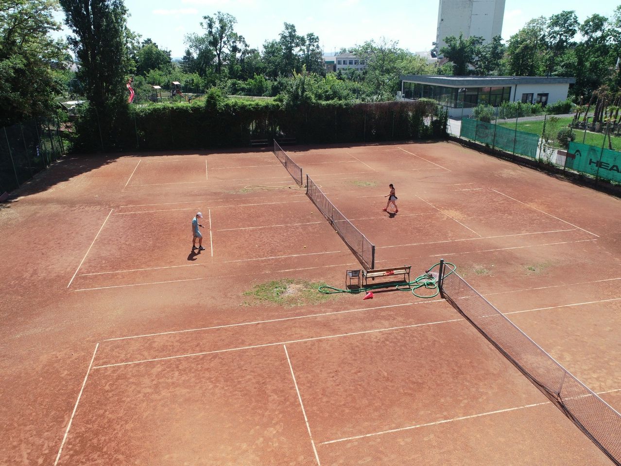 Eine Vogelperspektive eines Tennisplatzes mit zwei darauf stehenden Personen. Eine Person ist in der Nähe des Netzes, die andere geht. Der Platz hat weiße Linien und eine Bank.