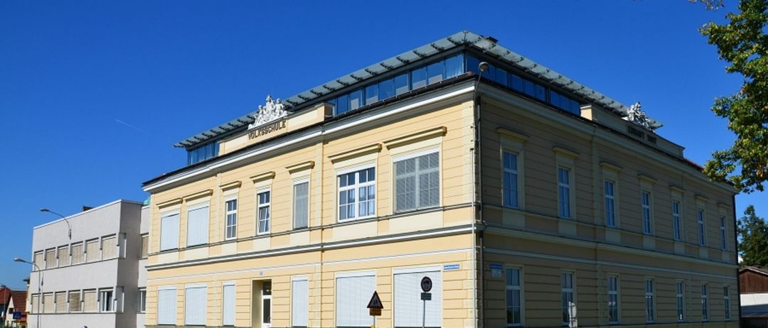 Ein gelbes Gebäude mit vielen Fenstern heißt Volksschule. Das Gebäude hat ein flaches Dach und steht an einem sonnigen Tag.