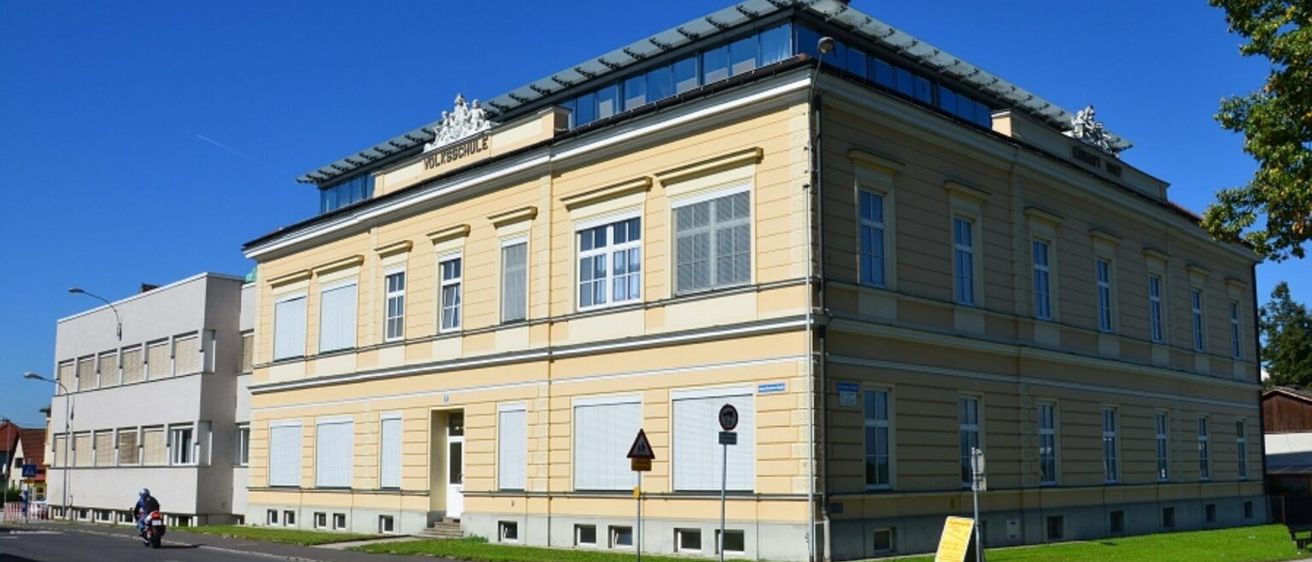 Ein gelbes Gebäude mit mehreren Fenstern und einem Schild mit der Aufschrift 'Volksschule' oben. Das Gebäude hat ein modernes Design mit einer glasüberdachten Terrasse. Es befindet sich an einer Straße mit Gras und einem Verkehrszeichen davor.