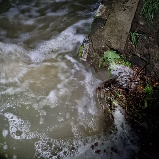 Eine Nahaufnahme eines Flusses mit turbulentem Wasser, das über eine Betonkante fließt. Das Wasser hat eine bräunliche Farbe und spritzt mit Blasen.