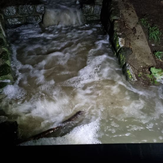 Eine Tiefwinkelaufnahme zeigt turbulentes Wasser, das durch einen Steinkanal fließt. Das Wasser ist schmutzig, mit Schaum auf der Oberfläche. Ein großer Baumstamm liegt im Kanal.