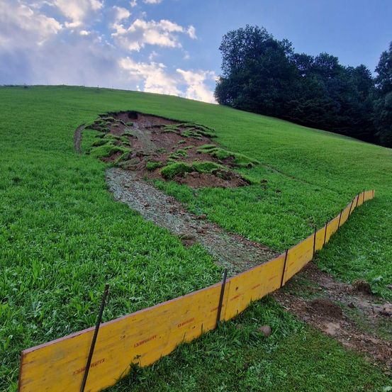 Ein grüner Grashügel hat ein großes Loch und einen kleinen Pfad. Ein gelber Zaun verläuft entlang der rechten Seite des Hügels.