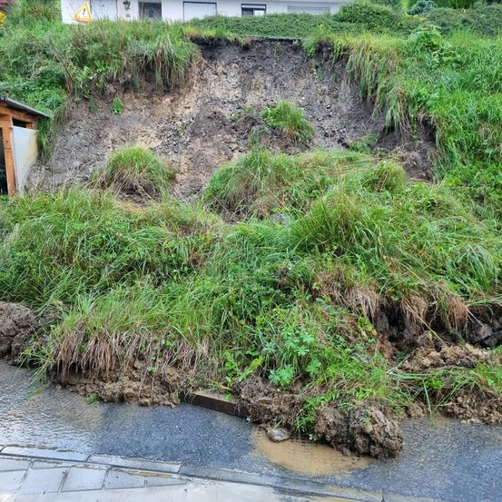 Ein Hang mit einem Erdrutsch zeigt freiliegende Erde und gestörtes Gras. Links ist ein kleiner Schuppen zu sehen.