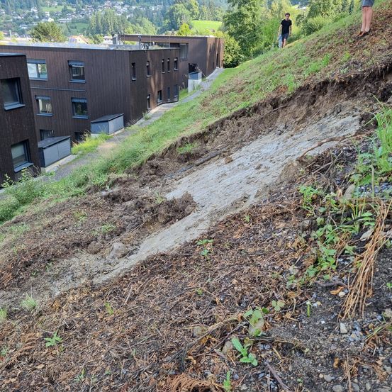 Ein Hang mit einem Erdrutsch zeigt einen beschädigten Hang mit erodiertem Boden und Schutt. Dahinter steht ein modernes Mehrfamilienhaus mit Glasfenstern. Zwei Personen stehen am Hang, eine hält eine Flasche.