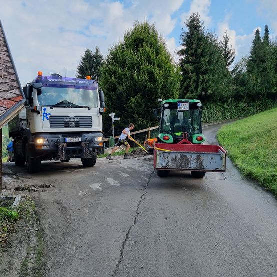 Ein weißer Lkw und ein grüner Traktor sind auf einer Straße geparkt, mit einem Mann, der in der Mitte einen Besen benutzt.