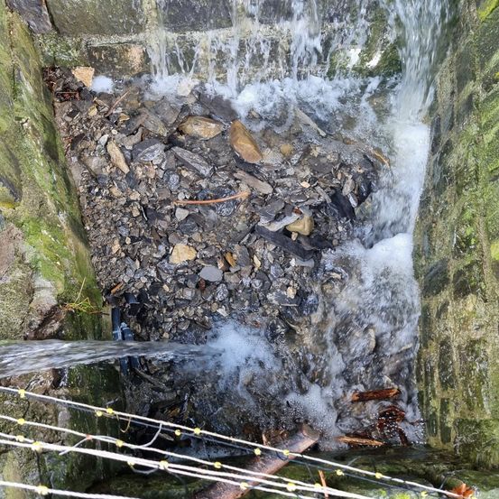 Wasser fließt aus einem Betonkanal und füllt ein Becken mit Steinen und Schutt. Die Steine sind mit Moos überzogen.