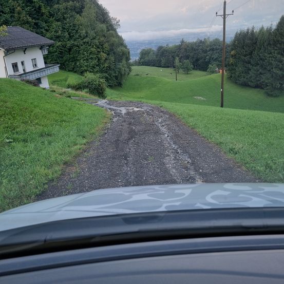 Eine Schotterstraße mit einem Haus auf der linken Seite und einer Fernsicht auf ein Tal und einen Strommast.