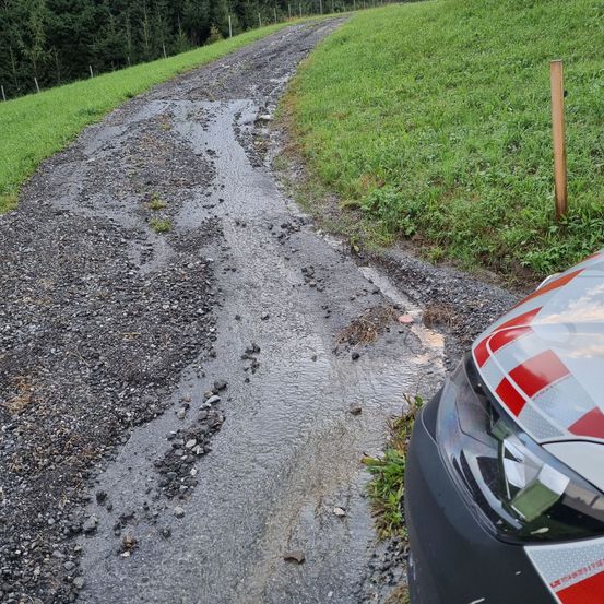 Eine Nahaufnahme einer beschädigten Straße mit einem geparkten Auto in der Nähe. Die Straße ist schlammig mit Schutt und einer flachen Wasserpfütze.