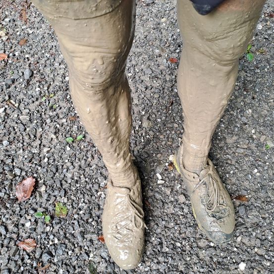 Die Beine einer Person sind mit Schlamm bedeckt und stehen auf einer felsigen Oberfläche. Die Stiefel und Schuhe sind stark verschmutzt.