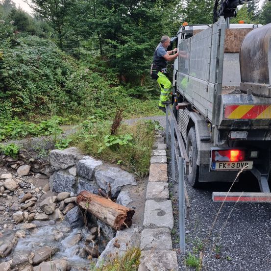 Ein Mann klettert auf einen Lastwagen, der an einer Straße mit fließendem Wasser und Steinen geparkt ist.