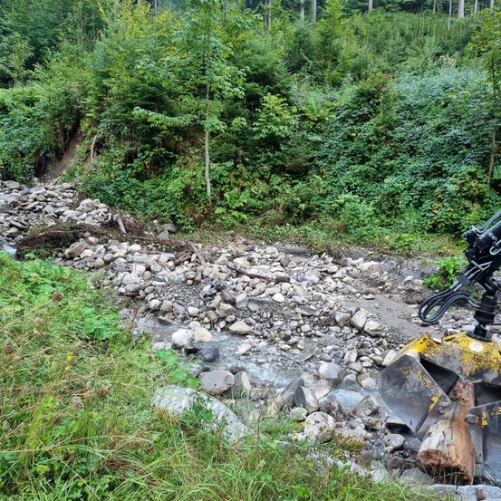 Ein Wasserstrom fließt durch eine felsige Landschaft mit einem großen gelben Bagger auf der rechten Seite. Das Gebiet ist von üppiger Grünfläche umgeben.