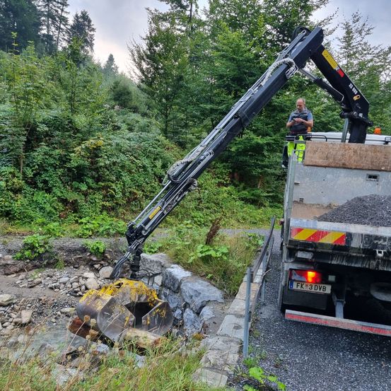 Ein Mann steht auf einem Lastwagen und beaufsichtigt den Einsatz eines Krans, der schwere Materialien hebt. Der Kran befindet sich am Straßenrand, in der Nähe einer Steinmauer und eines Gewässers. Das umliegende Gebiet ist von üppigem Baum- und Pflanzenbestand geprägt.