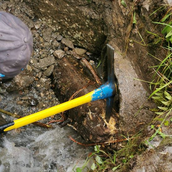 Eine Person in einem grauen Hut benutzt eine gelbe und blaue Spitzhacke, um in einem felsigen Bereich am Bach zu graben.