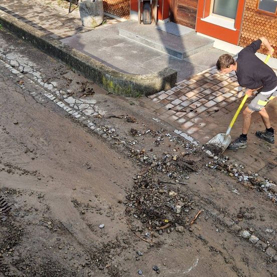 Ein Mann reinigt eine Straße mit einem Spaten und entfernt Schmutz und Ablagerungen. Die Straße wird von einem gepflasterten Gehweg und einem Gebäude mit einer Treppe gesäumt.