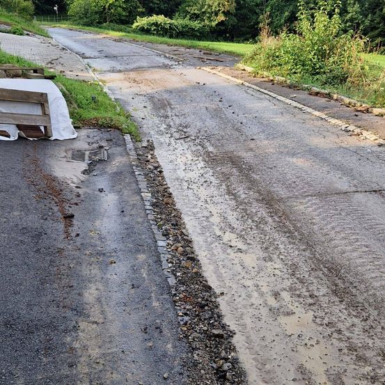 Eine beschädigte Straße mit einer Wasserpfütze und Kies auf der Seite. Eine Holzpalette befindet sich auf der linken Seite.