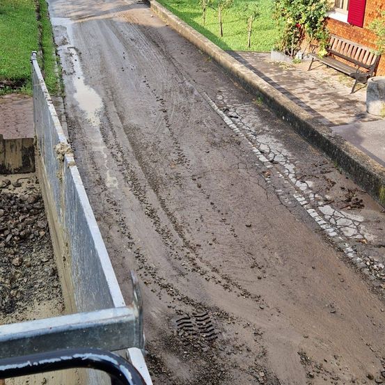 Eine schmutzig Straße mit Reifenspuren, begrenzt durch einen Betonrand und einen gepflasterten Gehweg. Eine Bank und eine Pflanze befinden sich auf dem Gehweg neben einem Gebäude mit roten Fensterläden.