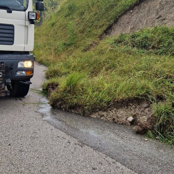 Ein Lkw mit eingeschalteten Scheinwerfern steht auf einer Straße, nahe einem Grashang mit etwas freiliegender Erde.