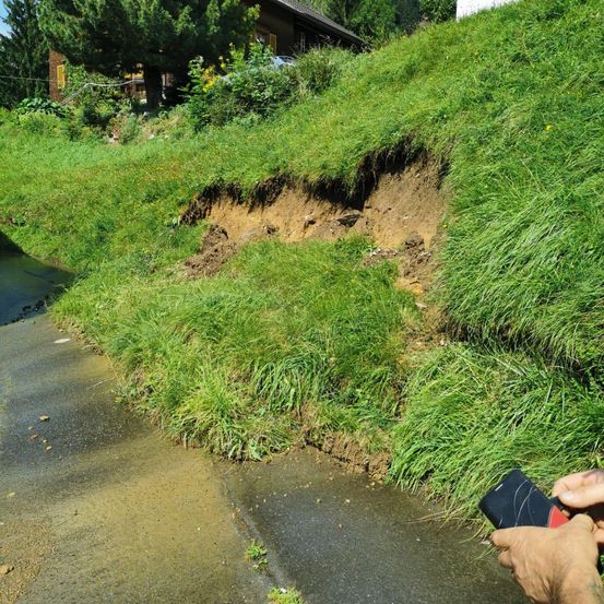 Eine Person hält ein Gerät auf einer nassen Straße in der Nähe einer Böschung mit Gras und einem Haus.